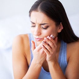 A young woman sitting on her bed and holding her cheek in pain and needing root canal therapy in Arlington