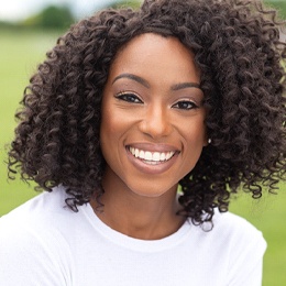 A young woman wearing a white blouse, sitting outside, and smiling after seeing her cosmetic dentist in Arlington about porcelain veneers