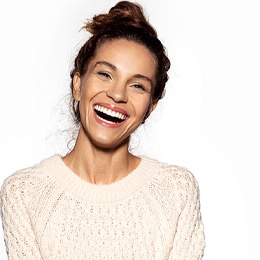 A young woman wearing a cream-colored sweater and smiling, showing off her new and improved smile after porcelain veneers