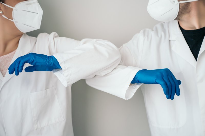 two medical professionals wearing lab coats, gloves, and masks performing an elbow bump