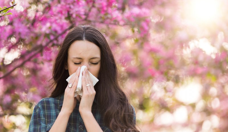 a woman stands outside during the spring and blows her nose because of seasonal allergies