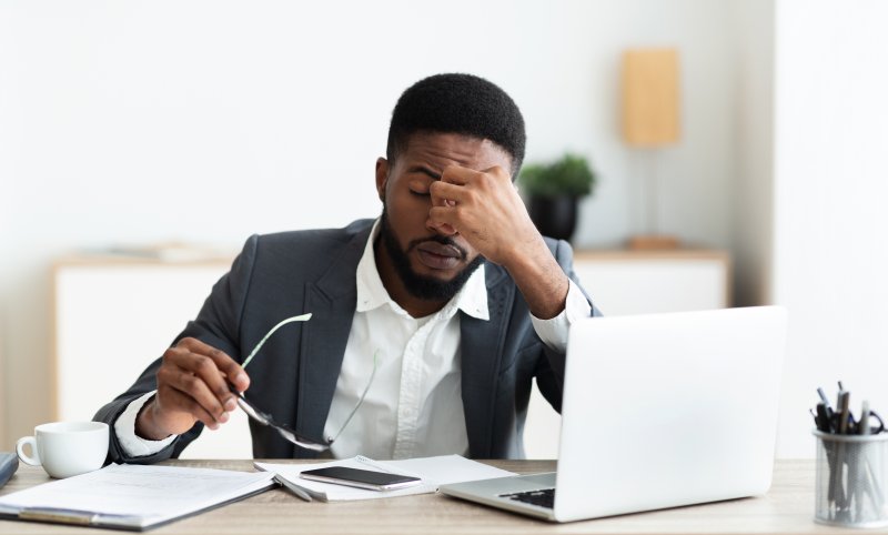 a young man seated at his desk and holding the area between his eyes because of stress